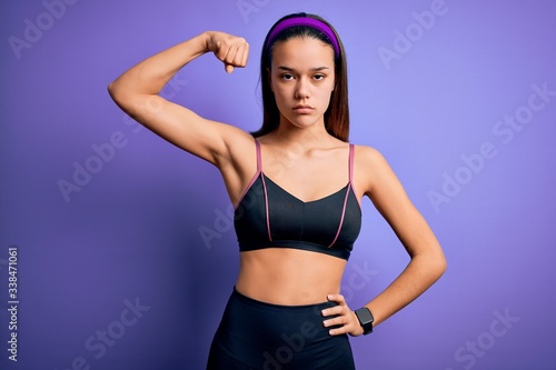Young beautiful sporty girl doing sport wearing sportswear over isolated purple background Strong person showing arm muscle, confident and proud of power © Krakenimages.com