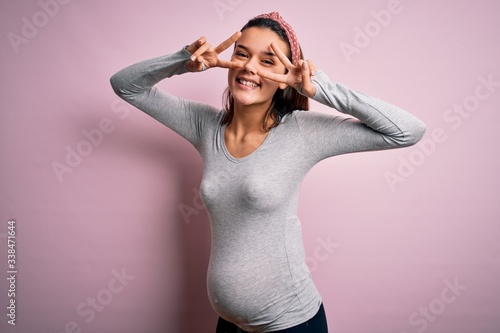 Young beautiful teenager girl pregnant expecting baby over isolated pink background Doing peace symbol with fingers over face, smiling cheerful showing victory photo