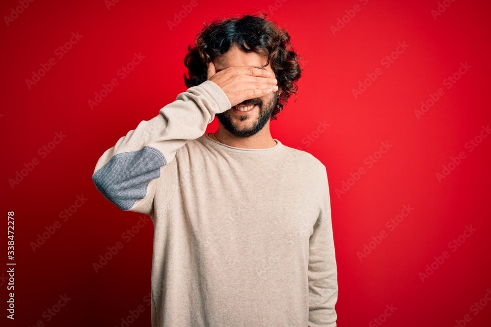 Young handsome man with beard wearing casual sweater standing over red background smiling and laughing with hand on face covering eyes for surprise. Blind concept. Young handsome man with beard wearing casual sweater standing over red background smiling and laughing with hand on face covering eyes for surprise. Blind concept.