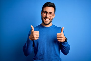 Young handsome man with beard wearing casual sweater and glasses over blue background success sign doing positive gesture with hand, thumbs up smiling and happy. Cheerful expression and winner