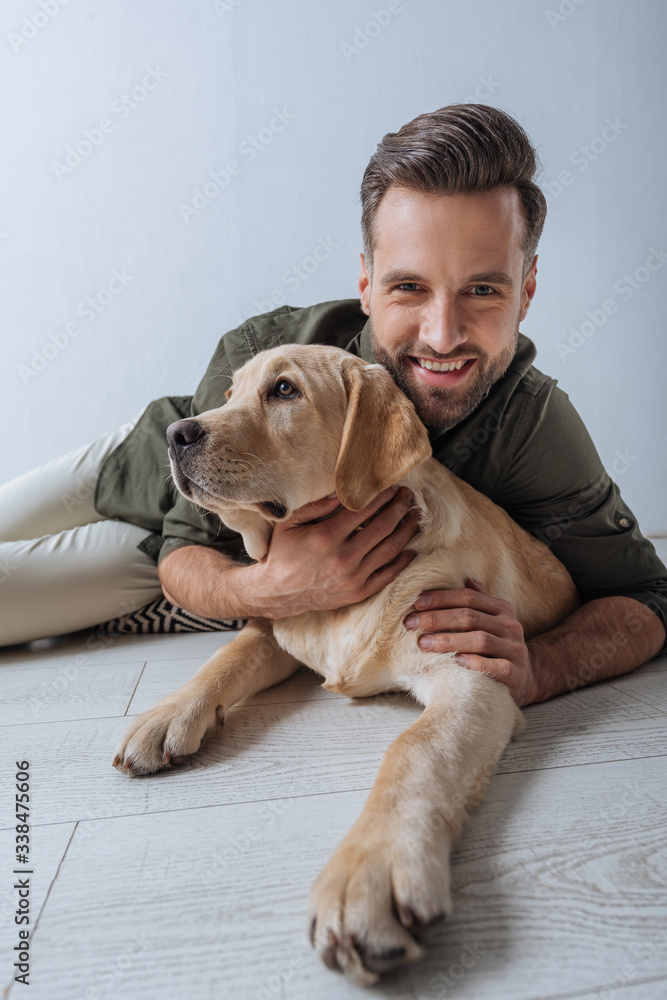 Selective focus of man smiling at camera while petting golden retriever on floor on grey background Selective focus of man smiling at camera while petting golden retriever on floor on grey background