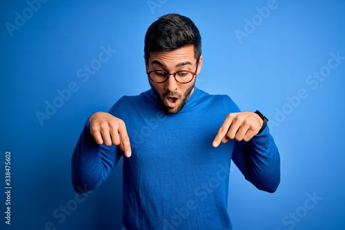 Obraz na plátně Young handsome man with beard wearing casual sweater and glasses over blue backg