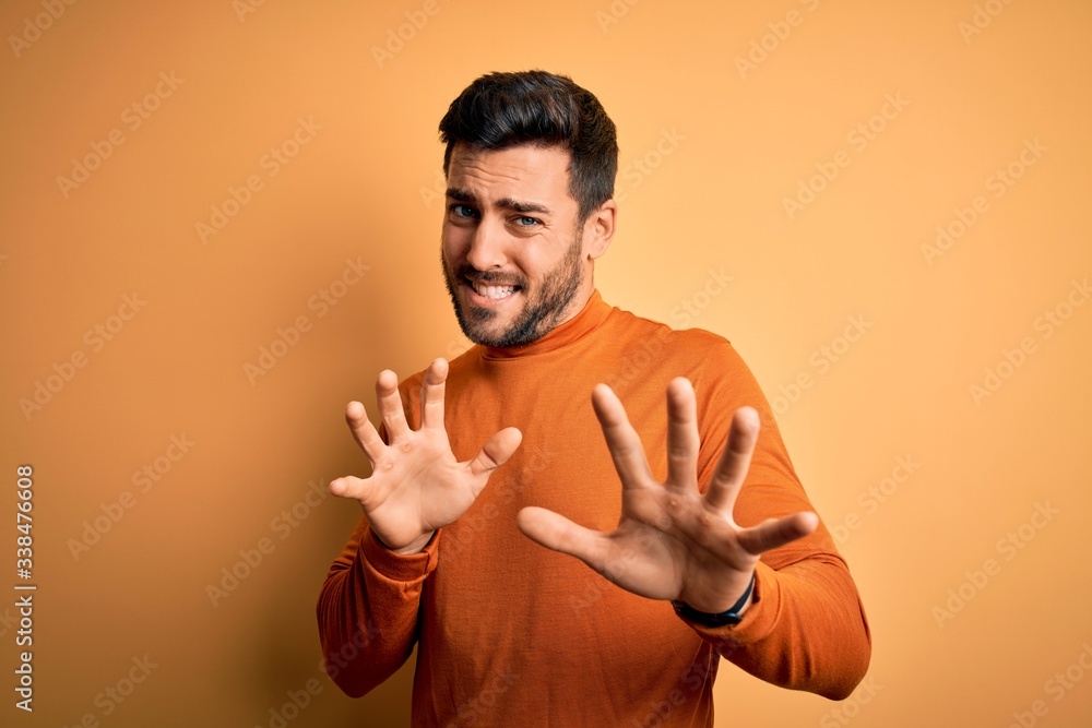 Young handsome man with beard wearing casual sweater standing over yellow background disgusted expression, displeased and fearful doing disgust face because aversion reaction. With hands raised Young handsome man with beard wearing casual sweater standing over yellow background disgusted expression, displeased and fearful doing disgust face because aversion reaction. With hands raised