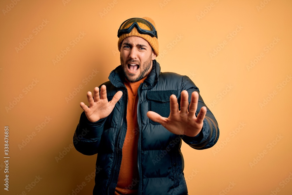 Young handsome skier man with beard wearing snow sportswear and ski goggles afraid and terrified with fear expression stop gesture with hands, shouting in shock. Panic concept. Young handsome skier man with beard wearing snow sportswear and ski goggles afraid and terrified with fear expression stop gesture with hands, shouting in shock. Panic concept.