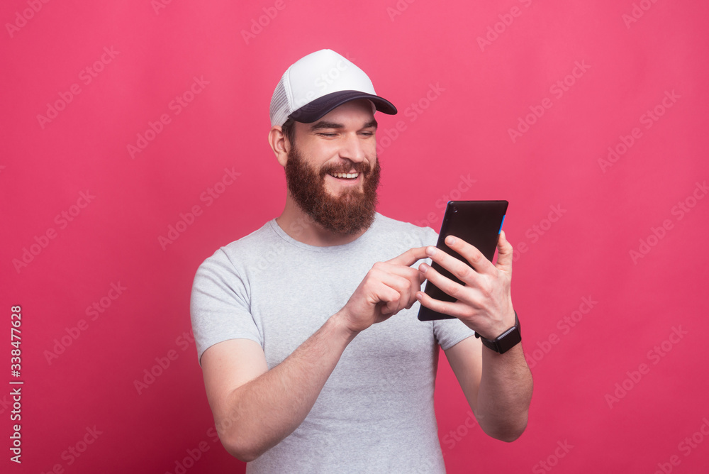 Portrait of cheerful man with beard smiling and using his tablet over pink background Portrait of cheerful man with beard smiling and using his tablet over pink background