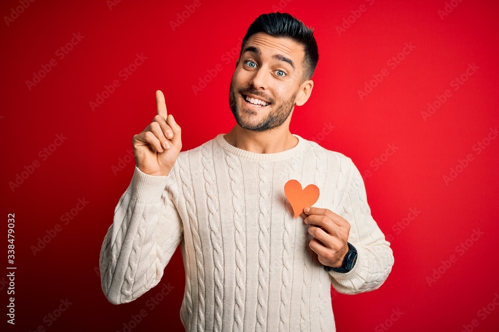 Young handsome man holding red shape heart as romantic and health symbol surprised with an idea or question pointing finger with happy face, number one Young handsome man holding red shape heart as romantic and health symbol surprised with an idea or question pointing finger with happy face, number one