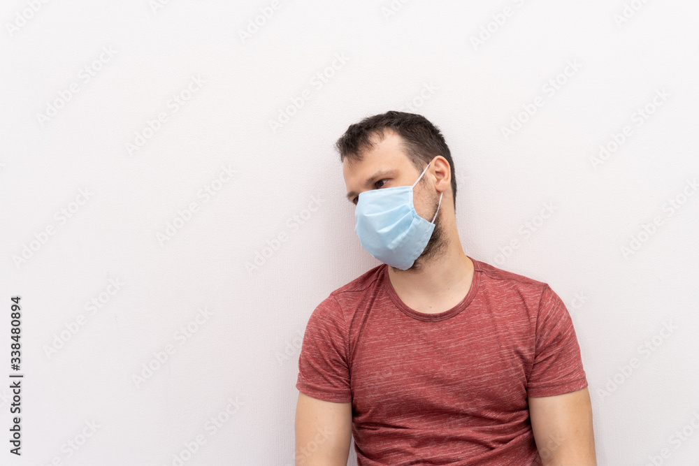 Caucasian man in facial surgical medical protective mask stay near wall, he is tired of coronavirus, staying home and feels sick, waits for the end of quarantine. Caucasian man in facial surgical medical protective mask stay near wall, he is tired of coronavirus, staying home and feels sick, waits for the end of quarantine.