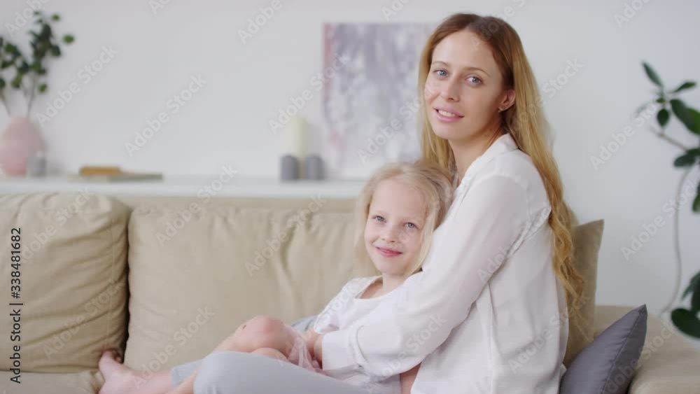 Portrait shot of happy young woman and her cute little daughter with blond hair sitting on couch and hugging while looking at camera Portrait shot of happy young woman and her cute little daughter with blond hair sitting on couch and hugging while looking at camera