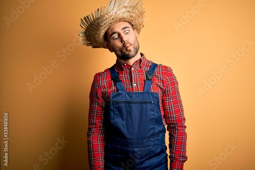 Young rural farmer man wearing bib overall and countryside hat over yellow background making fish face with lips, crazy and comical gesture. Funny expression.