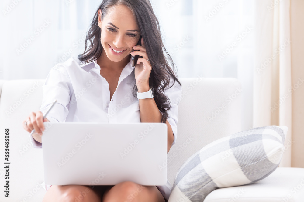 Beautiful girl looking into her laptop on her knees, smiling and holding a pen in the right hand and a phone on her ear with the left hand, sitting on a sofa in her light lit apartment