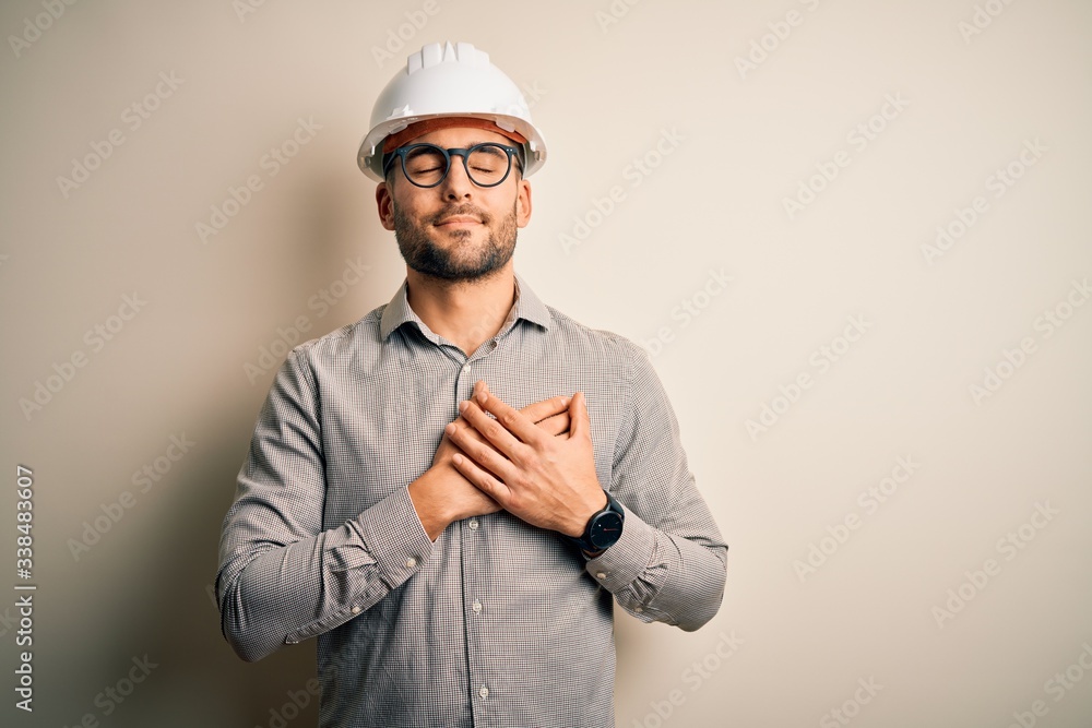 Young architect man wearing builder safety helmet over isolated background smiling with hands on chest with closed eyes and grateful gesture on face. Health concept.