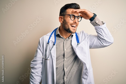 Young doctor man wearing glasses, medical white robe and stethoscope over isolated background very happy and smiling looking far away with hand over head. Searching concept.