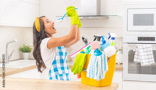 Housekeeping young lady with a yellow bucket full of cleaning stuff on her kitchen desk doing frolics with a kitchen brush pretending to sing into it