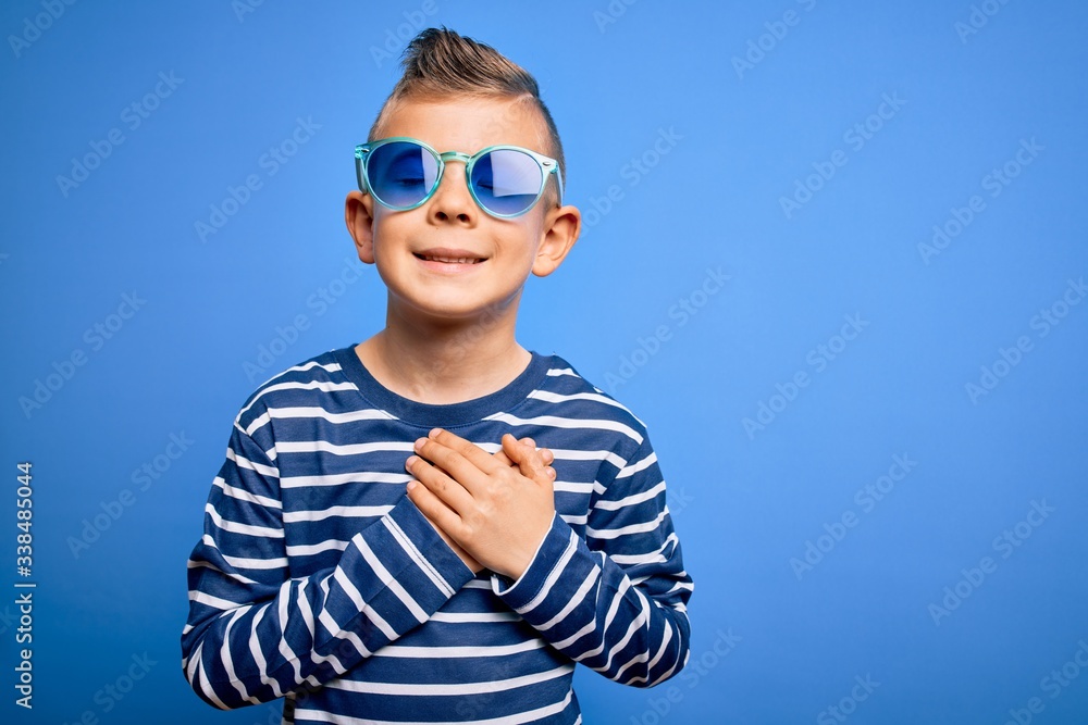 Young little caucasian kid with blue eyes standing wearing sunglasses over blue background smiling with hands on chest with closed eyes and grateful gesture on face. Health concept. Young little caucasian kid with blue eyes standing wearing sunglasses over blue background smiling with hands on chest with closed eyes and grateful gesture on face. Health concept.