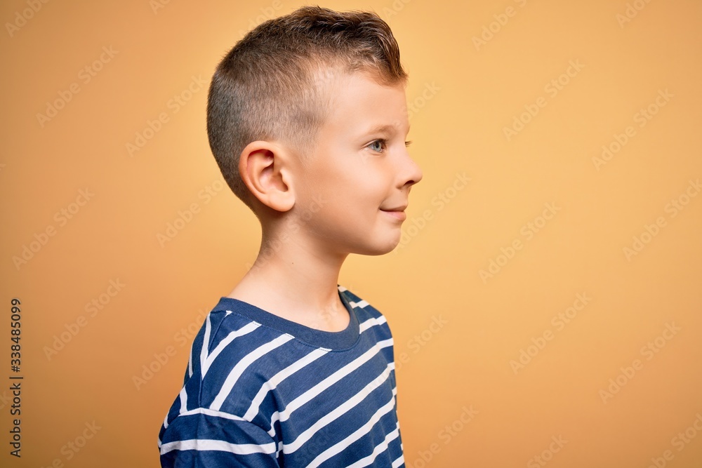 Young little caucasian kid with blue eyes wearing nautical striped shirt over yellow background looking to side, relax profile pose with natural face with confident smile. Young little caucasian kid with blue eyes wearing nautical striped shirt over yellow background looking to side, relax profile pose with natural face with confident smile.