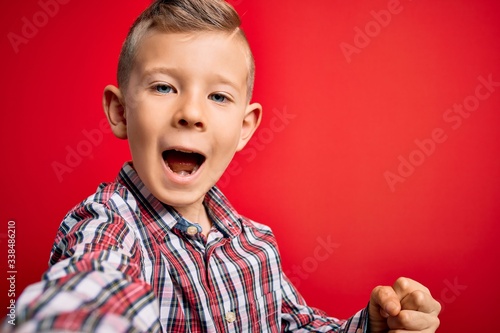 Close up of young little caucasian kid with blue eyes taking a selfie photo over red background screaming proud and celebrating victory and success very excited, cheering emotion