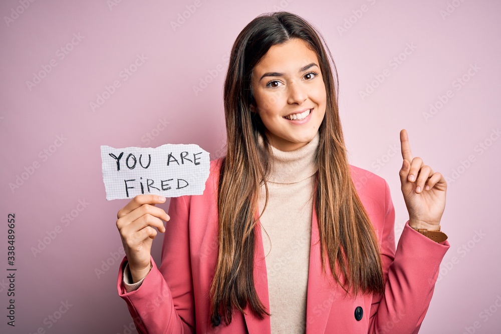 Young beautiful girl holding paper with you are fired message over isolated pink background surprised with an idea or question pointing finger with happy face, number one Young beautiful girl holding paper with you are fired message over isolated pink background surprised with an idea or question pointing finger with happy face, number one