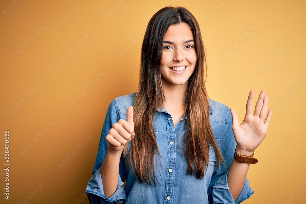 Young beautiful girl wearing casual denim shirt standing over isolated yellow background showing and pointing up with fingers number six while smiling confident and happy. Young beautiful girl wearing casual denim shirt standing over isolated yellow background showing and pointing up with fingers number six while smiling confident and happy.