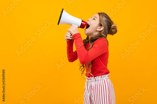 european teenager girl screaming news in a loudspeaker and stands sideways on a yellow studio background with copy space