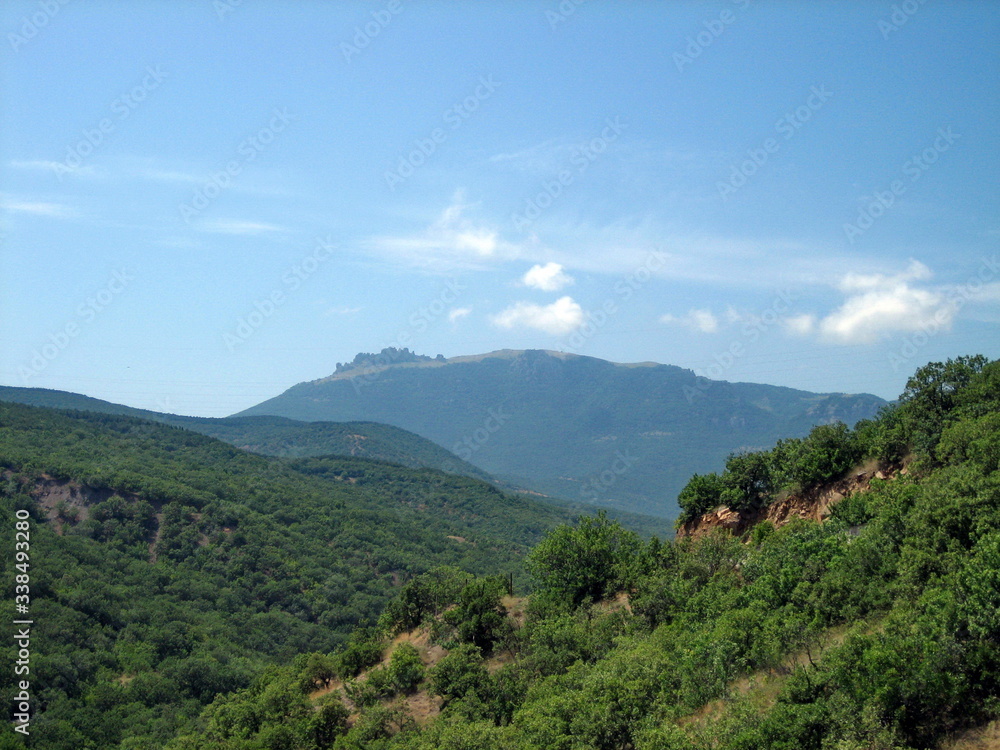 Valley of hills covered with low green forest. In the distance, the silhouettes of high mountains turn blue. Clear blue sky with small clouds.