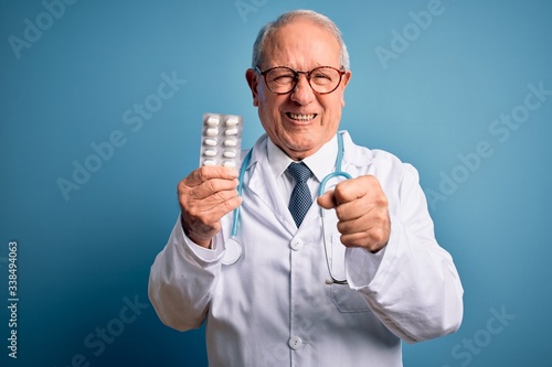 Senior grey haired doctor man holding pharmaceutical pills over blue background annoyed and frustrated shouting with anger, crazy and yelling with raised hand, anger concept