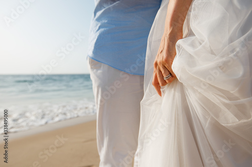 Newlyweds honeymoon. Bride holding her white dress by the hand with wedding rings. Couple on the ocean beach