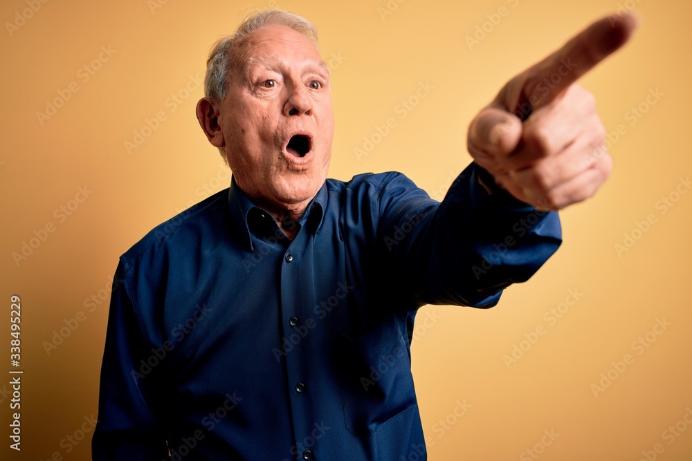 Grey haired senior man wearing casual blue shirt standing over yellow background Pointing with finger surprised ahead, open mouth amazed expression, something on the front Grey haired senior man wearing casual blue shirt standing over yellow background Pointing with finger surprised ahead, open mouth amazed expression, something on the front