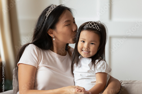 Asian young mother kisses little cutie daughter sitting on couch in living room wearing tiara crown accessory spend funny time together at home. Concept of affection, family bonds care and protection