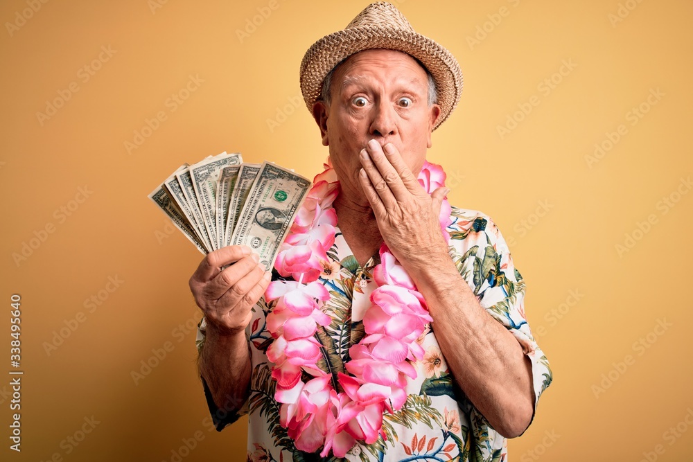 Grey haired senior man wearing summer hat and hawaiian lei holding bunch of dollars cover mouth with hand shocked with shame for mistake, expression of fear, scared in silence, secret concept Grey haired senior man wearing summer hat and hawaiian lei holding bunch of dollars cover mouth with hand shocked with shame for mistake, expression of fear, scared in silence, secret concept