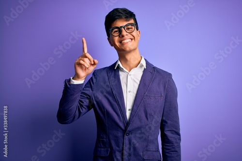 Young handsome business man wearing jacket and glasses over isolated purple background showing and pointing up with finger number one while smiling confident and happy.