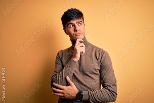 Young handsome man wearing casual turtleneck sweater over isolated yellow background with hand on chin thinking about question, pensive expression. Smiling with thoughtful face. Doubt concept. © Krakenimages.com