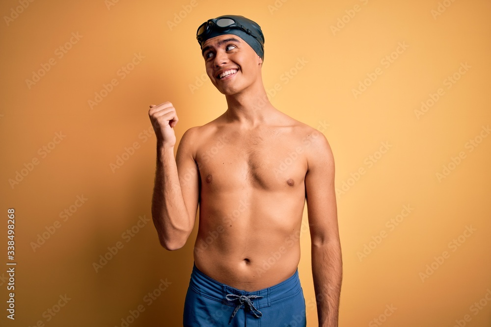 Young handsome man shirtless wearing swimsuit and swim cap over isolated yellow background smiling with happy face looking and pointing to the side with thumb up.