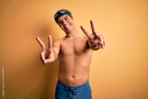 Young handsome man shirtless wearing swimsuit and swim cap over isolated yellow background smiling with tongue out showing fingers of both hands doing victory sign. Number two.