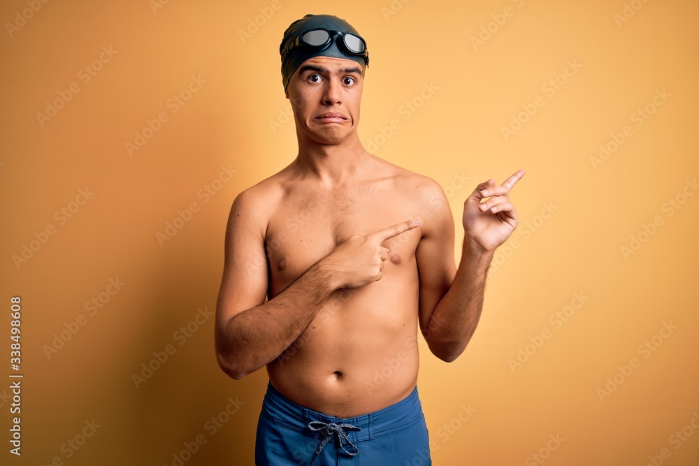 Young handsome man shirtless wearing swimsuit and swim cap over isolated yellow background Pointing aside worried and nervous with both hands, concerned and surprised expression