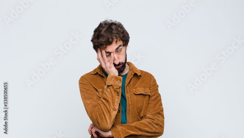 young handsome man feeling bored, frustrated and sleepy after a tiresome, dull and tedious task, holding face with hand against flat wall