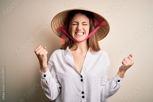 Young beautiful redhead woman wearing asian traditional conical hat over white background very happy and excited doing winner gesture with arms raised, smiling and screaming for success. Celebration