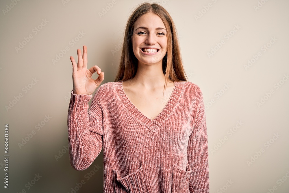 Young beautiful redhead woman wearing pink casual sweater over isolated white background smiling positive doing ok sign with hand and fingers. Successful expression. Young beautiful redhead woman wearing pink casual sweater over isolated white background smiling positive doing ok sign with hand and fingers. Successful expression.