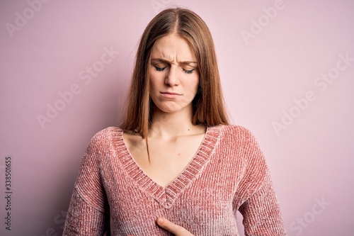 Young beautiful redhead woman wearing casual sweater over isolated pink background with hand on stomach because indigestion, painful illness feeling unwell. Ache concept. © Krakenimages.com