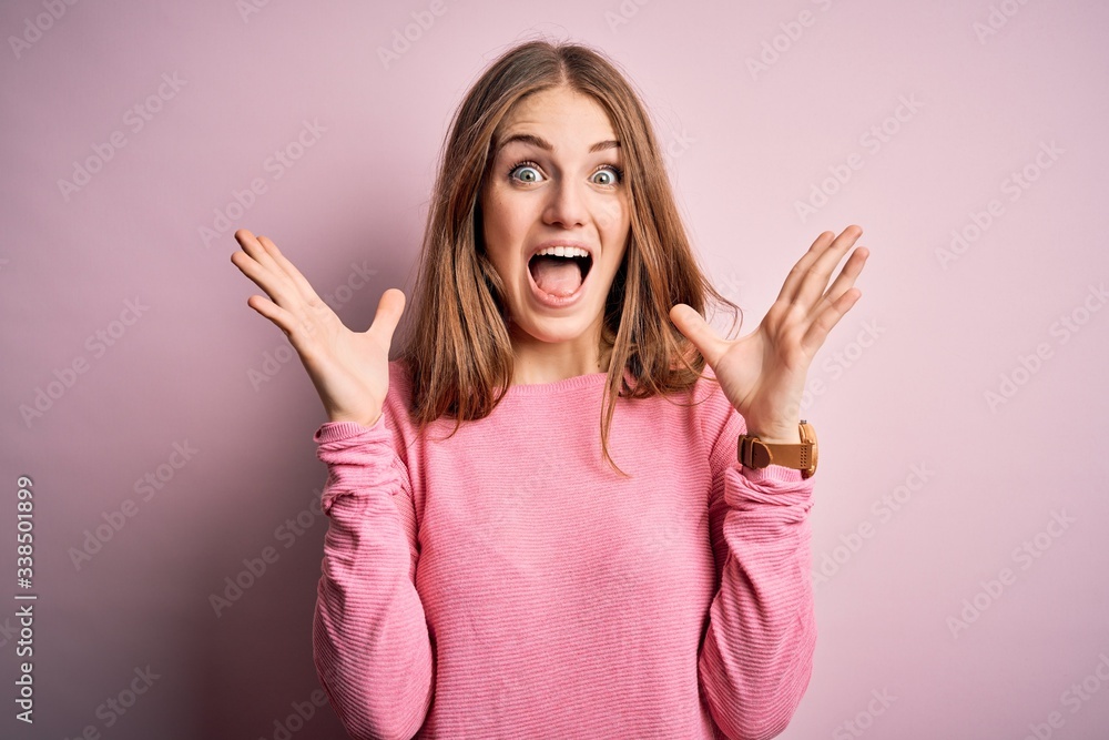 Young beautiful redhead woman wearing casual sweater over isolated pink background celebrating crazy and amazed for success with arms raised and open eyes screaming excited. Winner concept Young beautiful redhead woman wearing casual sweater over isolated pink background celebrating crazy and amazed for success with arms raised and open eyes screaming excited. Winner concept