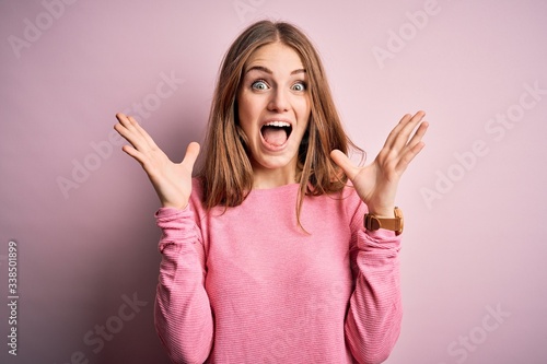 Young beautiful redhead woman wearing casual sweater over isolated pink background celebrating crazy and amazed for success with arms raised and open eyes screaming excited. Winner concept