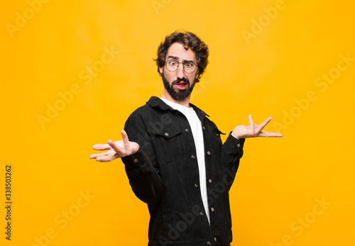 young crazy handsome man shrugging with a dumb, crazy, confused, puzzled expression, feeling annoyed and clueless against orange wall