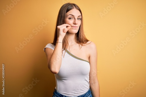 Young beautiful redhead woman wearing casual t-shirt over isolated yellow background mouth and lips shut as zip with fingers. Secret and silent  taboo talking