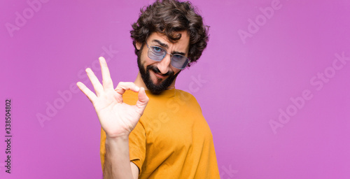 young crazy cool man feeling happy, relaxed and satisfied, showing approval with okay gesture, smiling against flat wall