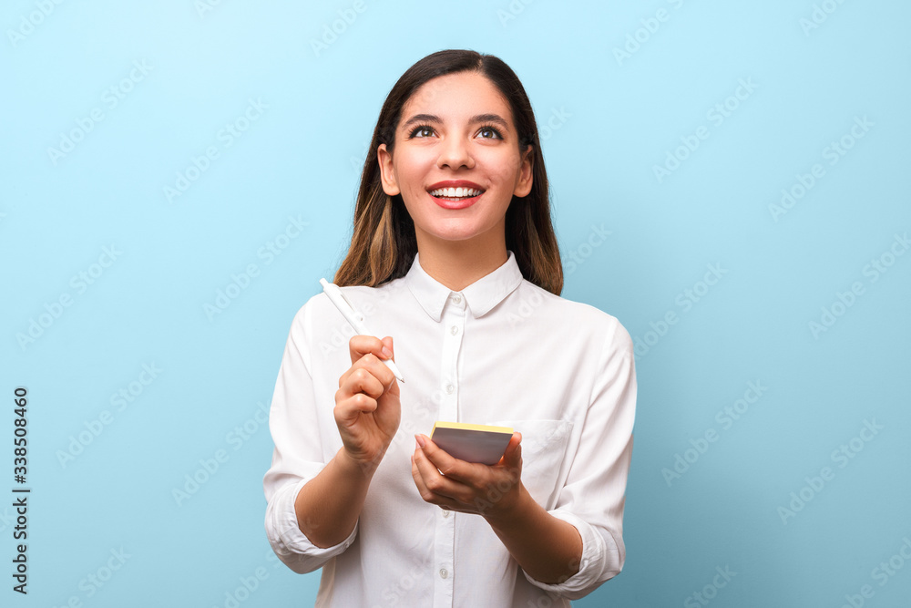 closeup young cheerful businesswoman with beautiful healthy smile holding pen making new ideas notes and interestedly looking up against blue background closeup young cheerful businesswoman with beautiful healthy smile holding pen making new ideas notes and interestedly looking up against blue background