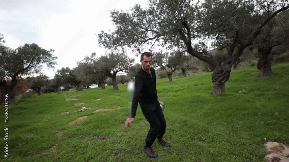 Juggling artist performing playful tricks with a club while holding 4 clubs in 1 hand. Slow motion wide shot with olive trees in the background. Juggling artist performing playful tricks with a club while holding 4 clubs in 1 hand. Slow motion wide shot with olive trees in the background.