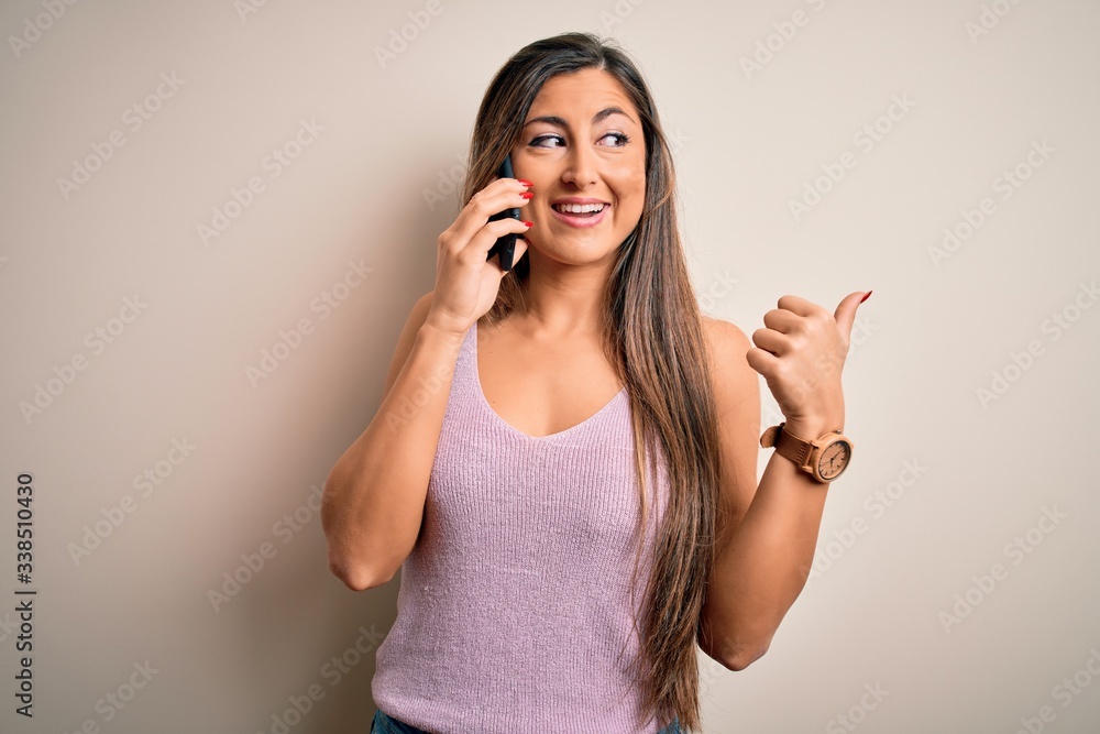 Young brunette woman having conversation talking on the smartphone over white background pointing and showing with thumb up to the side with happy face smiling Young brunette woman having conversation talking on the smartphone over white background pointing and showing with thumb up to the side with happy face smiling