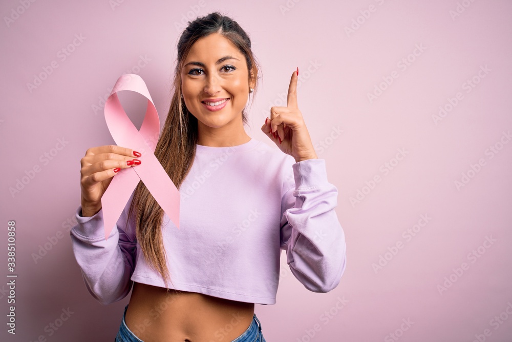 Young beautiful brunette woman holding pink cancer ribbon symbol over isolated background surprised with an idea or question pointing finger with happy face, number one Young beautiful brunette woman holding pink cancer ribbon symbol over isolated background surprised with an idea or question pointing finger with happy face, number one