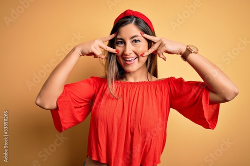 Young beautiful woman colorful summer style over yellow isolated background Doing peace symbol with fingers over face, smiling cheerful showing victory