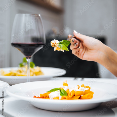 Female hand eating penne pasta with mozzarella cheeze with a glass of red wine in an italian restaurant, square format