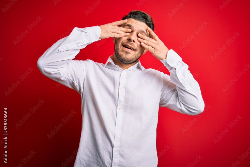 Young business man with blue eyes wearing elegant shirt standing over red isolated background covering eyes with hands smiling cheerful and funny. Blind concept. Young business man with blue eyes wearing elegant shirt standing over red isolated background covering eyes with hands smiling cheerful and funny. Blind concept.